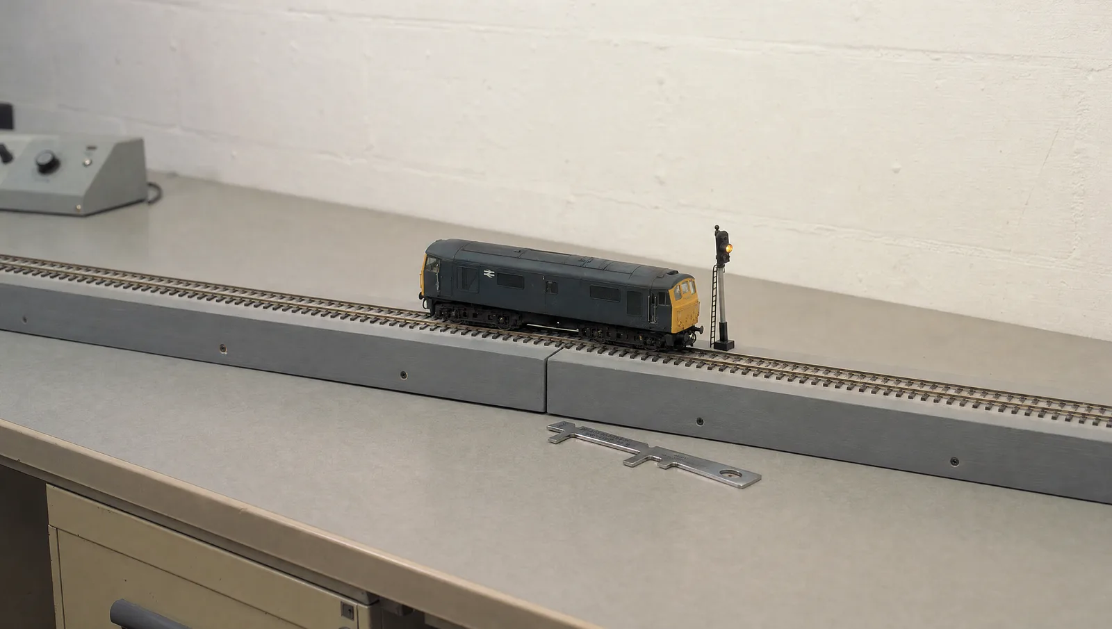 A small locomotive stands on the joint between two identical model railway baseboards on a 1980s test bench, a track gauge beside it and one amber signal lit.