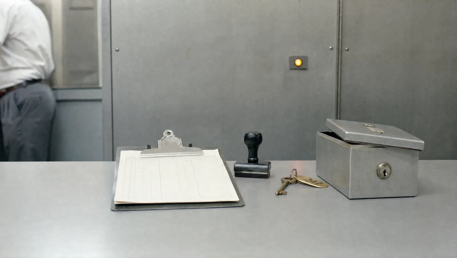 A checklist on a metal clipboard, a rubber stamp, a brass key and a small steel deposit box on a 1980s bank vault counter, with an amber lamp behind.